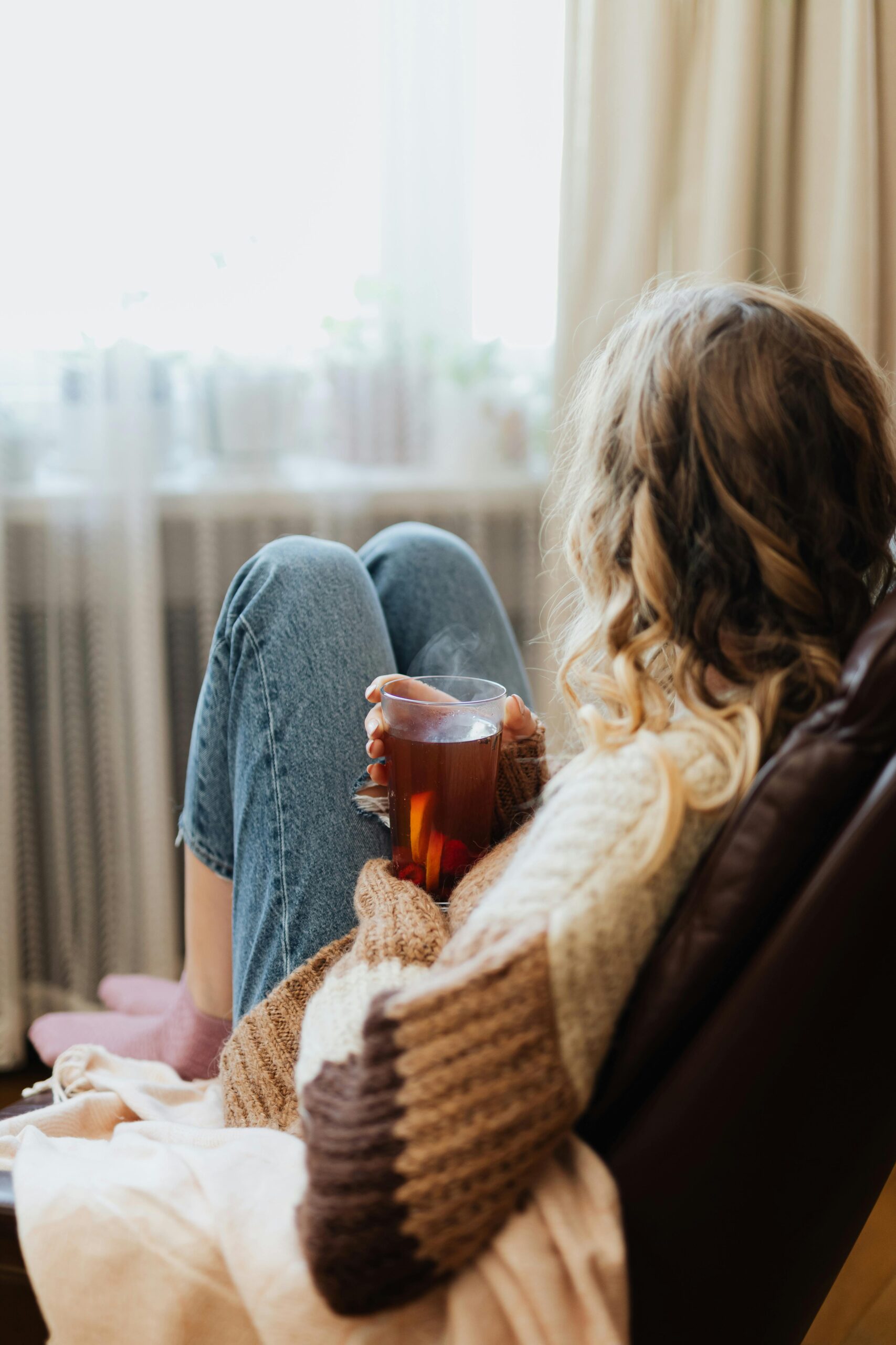 A woman sitting by a window in a sunlit room, holding a warm cup of tea and looking out thoughtfully; representing the emotional reflection during the IVF two-week wait.